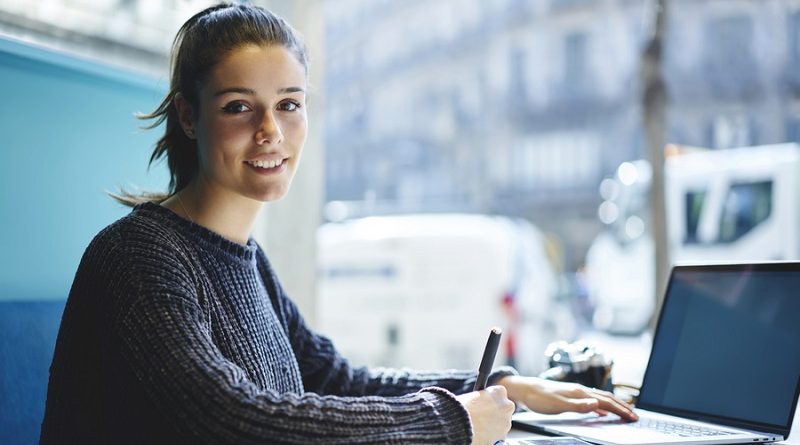 Portrait of charming young student sitting in coworking space looking at camera while making researches browsing information on laptop computer with mock up screen connected to wireless internet