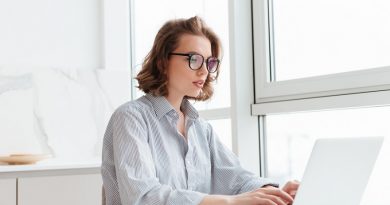 Photo of young concentrated woman in striped shirt using laptop while siting at table in light apartment