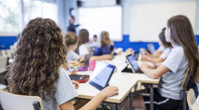 girl with brown curly hair studies in class with her tablet next to her classmates while they listen to the teacher