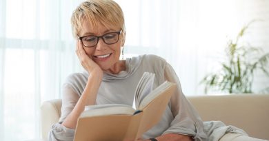 Senior blond woman in glasses resting on sofa and reading a book