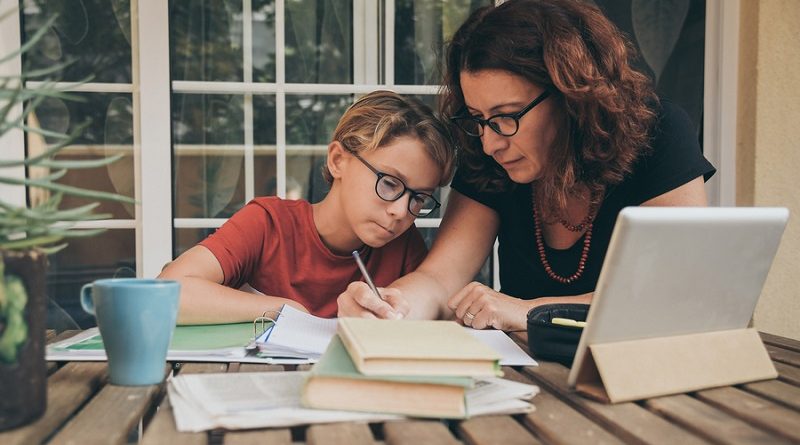 Young student doing homework at home with school books, newspaper and digital pad helped by his mother. Mum writing on the copybook teaching his son. Education, family lifestyle, homeschooling concept