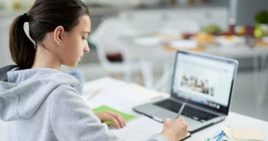 Concentrated latin teenage girl writing in her copybook while doing homework, using laptop, sitting at the table at home. Distance education, home schooling concept