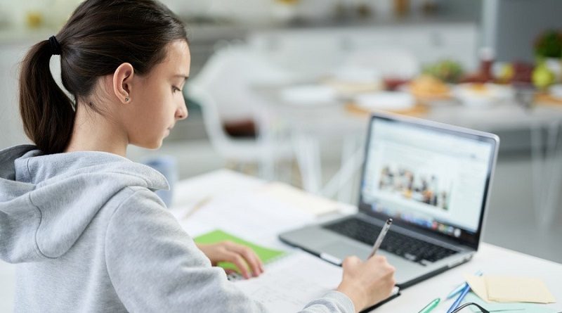 Concentrated latin teenage girl writing in her copybook while doing homework, using laptop, sitting at the table at home. Distance education, home schooling concept