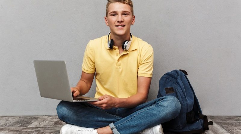 Portrait of a smiling casual teenage boy with backpack using laptop computer while sitting isolated over gray background