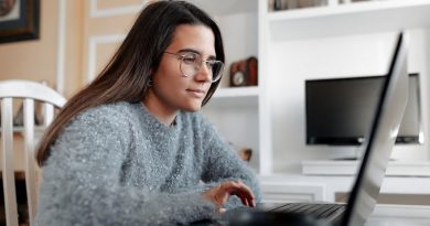 Young cheerful woman wearing a sweater in her home is sitting in her living room while browsing in the internet in the laptop.