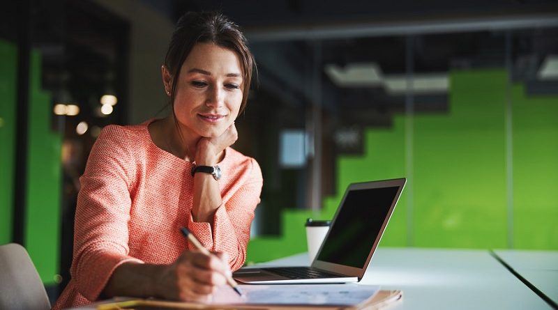 Elegant pretty Caucasian female business analyst writing something on the documents while sitting at the work desk in modern office space