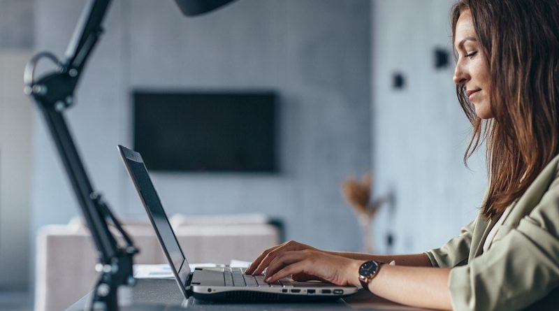 Woman works at home sitting at her desk with her laptop. Side view.