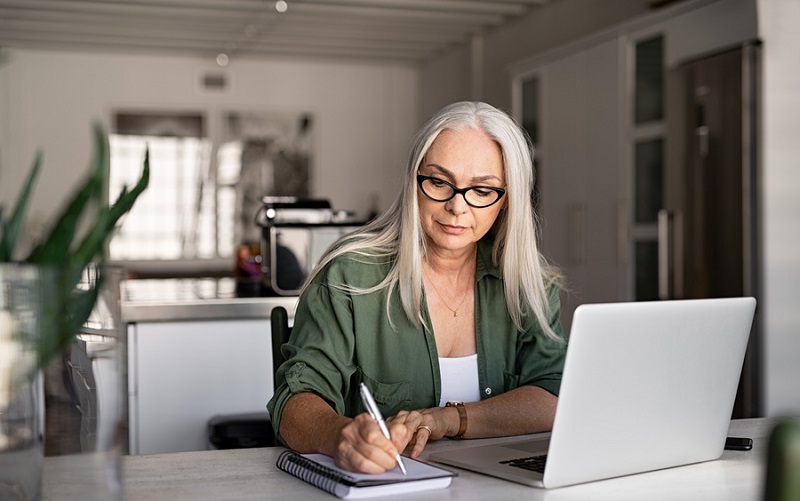 Senior stylish woman taking notes in notebook while using laptop at home. Old freelancer writing details on book while working on laptop in living room. Focused cool lady writing notary in notepad.